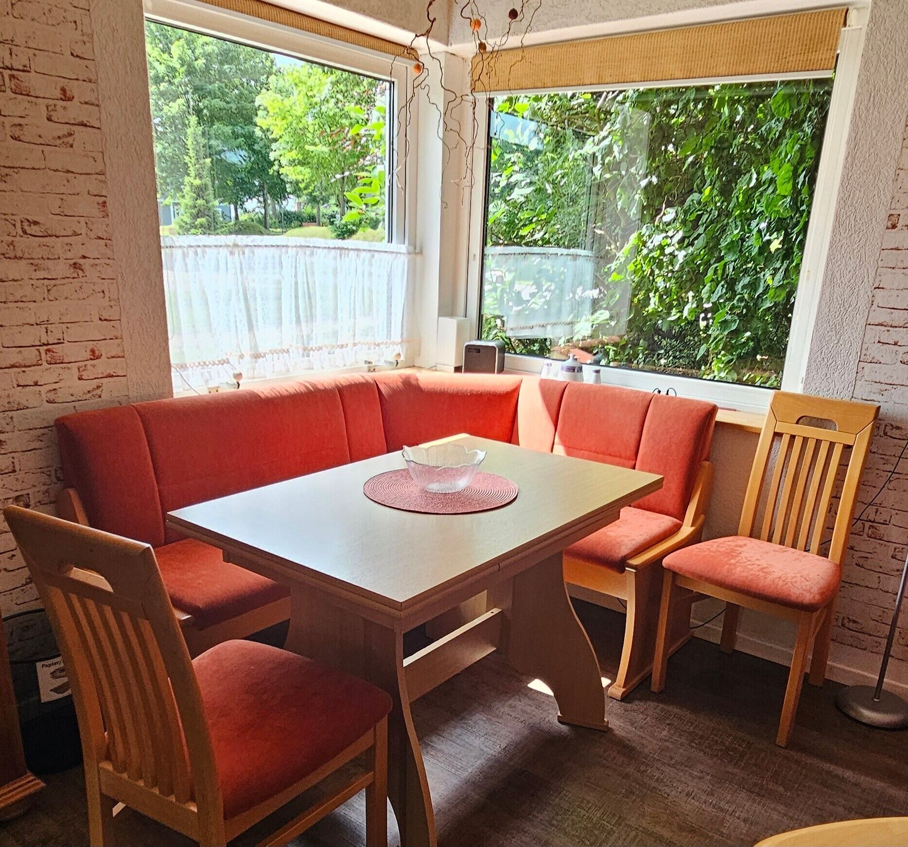 Een eetruimte met een rechthoekige tafel en rode fauteuils, een witte tafelkleed, en uitzicht op groen door grote ramen.