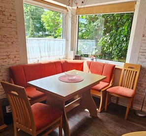 Comedor con mesa rectangular y sillas tapizadas rojas, mantel blanco y vista a zonas verdes a través de amplios ventanales.