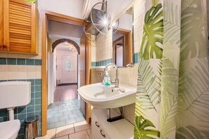 Un baño con lavamanos blanco, armarios de madera, un espejo y una cortina de ducha con estampado de hojas verdes.