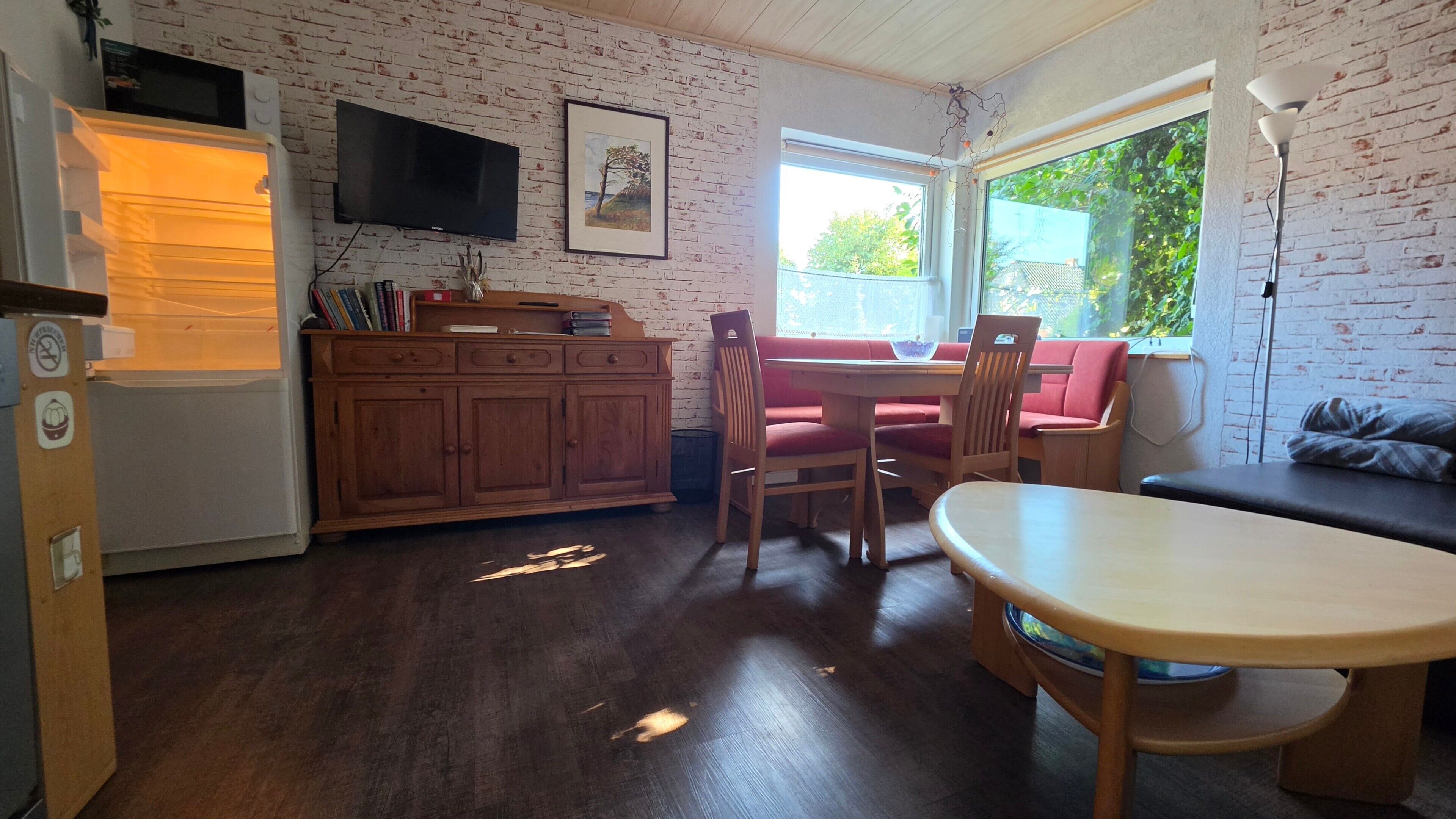 A kitchen with a wooden cabinet, a dining table with chairs, a television mounted on the wall, and a window with a view of greenery.