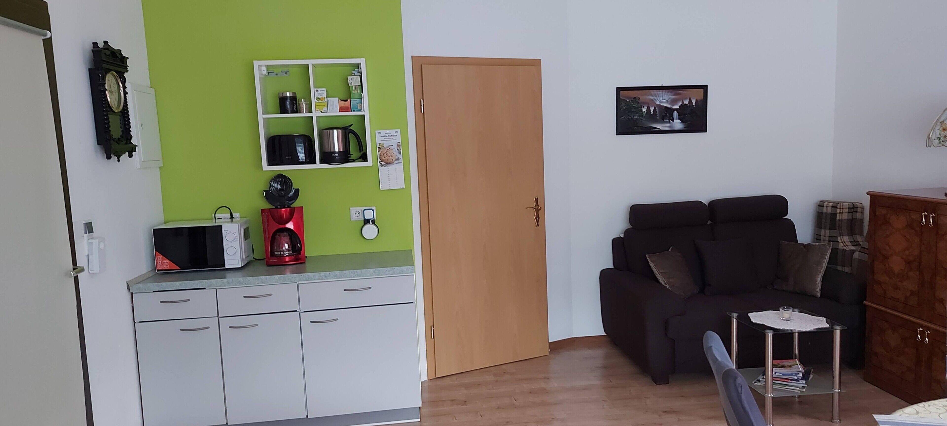 Une cuisine équipée d’un four à micro-ondes, d’une machine à café et d’une étagère contenant des ustensiles de cuisine. On y trouve également une porte en bois, un canapé noir et une petite table avec un plateau en verre.