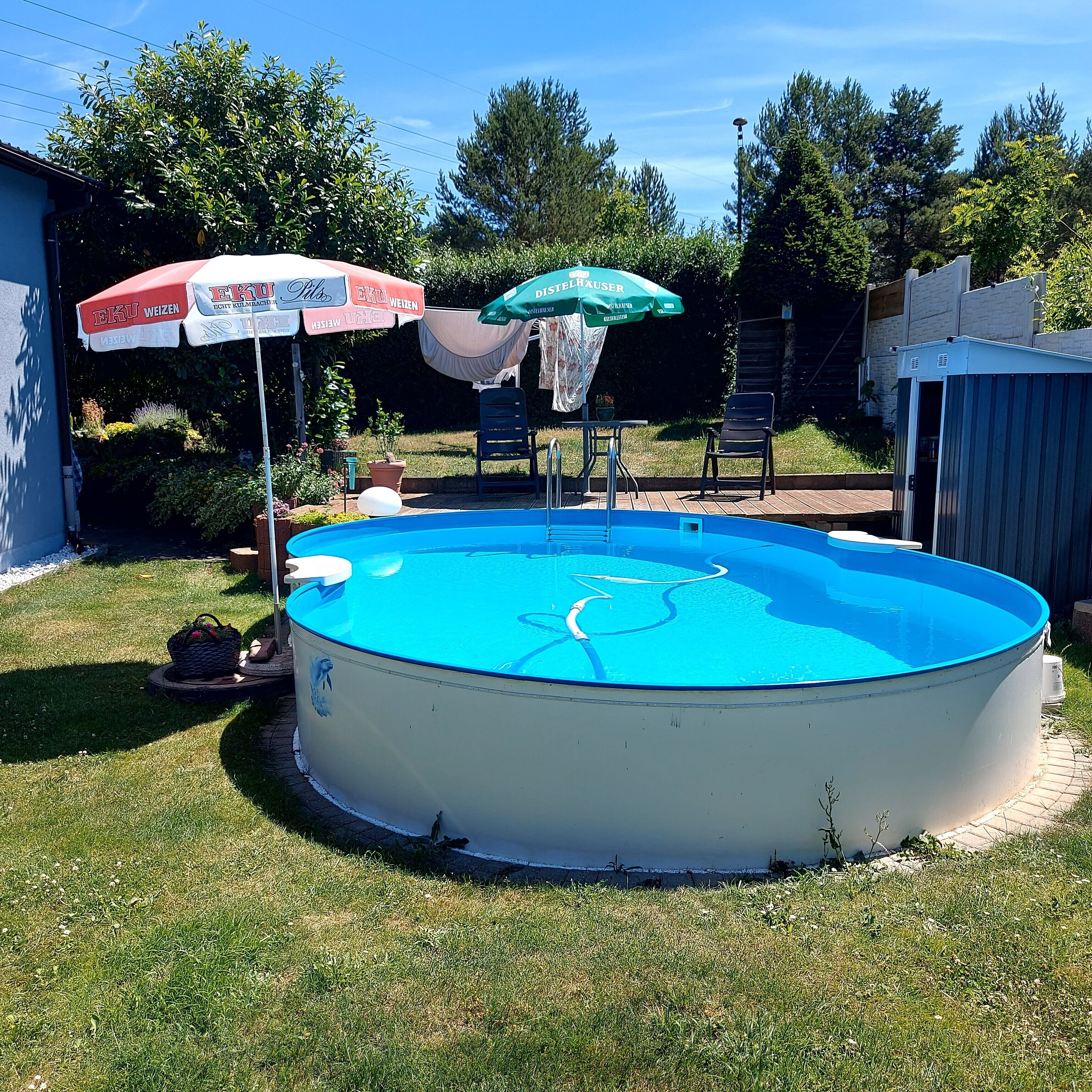 Une piscine ronde, installée à l’extérieur et hors sol, avec un intérieur bleu, entourée de pelouse, et deux parasols ainsi qu’une chaise visibles à l’arrière-plan.
