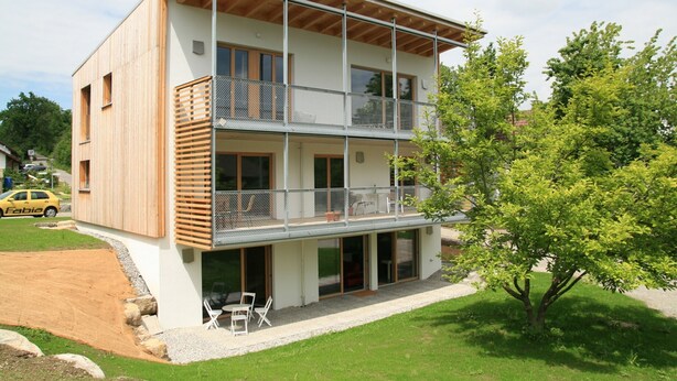 Ein modernes, zweistöckiges Gebäude mit Balkon, Auffahrt und Baum davor.