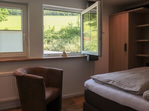 Quarto com cama, cadeira, guarda-roupa e janela com vista aberta para o verde.