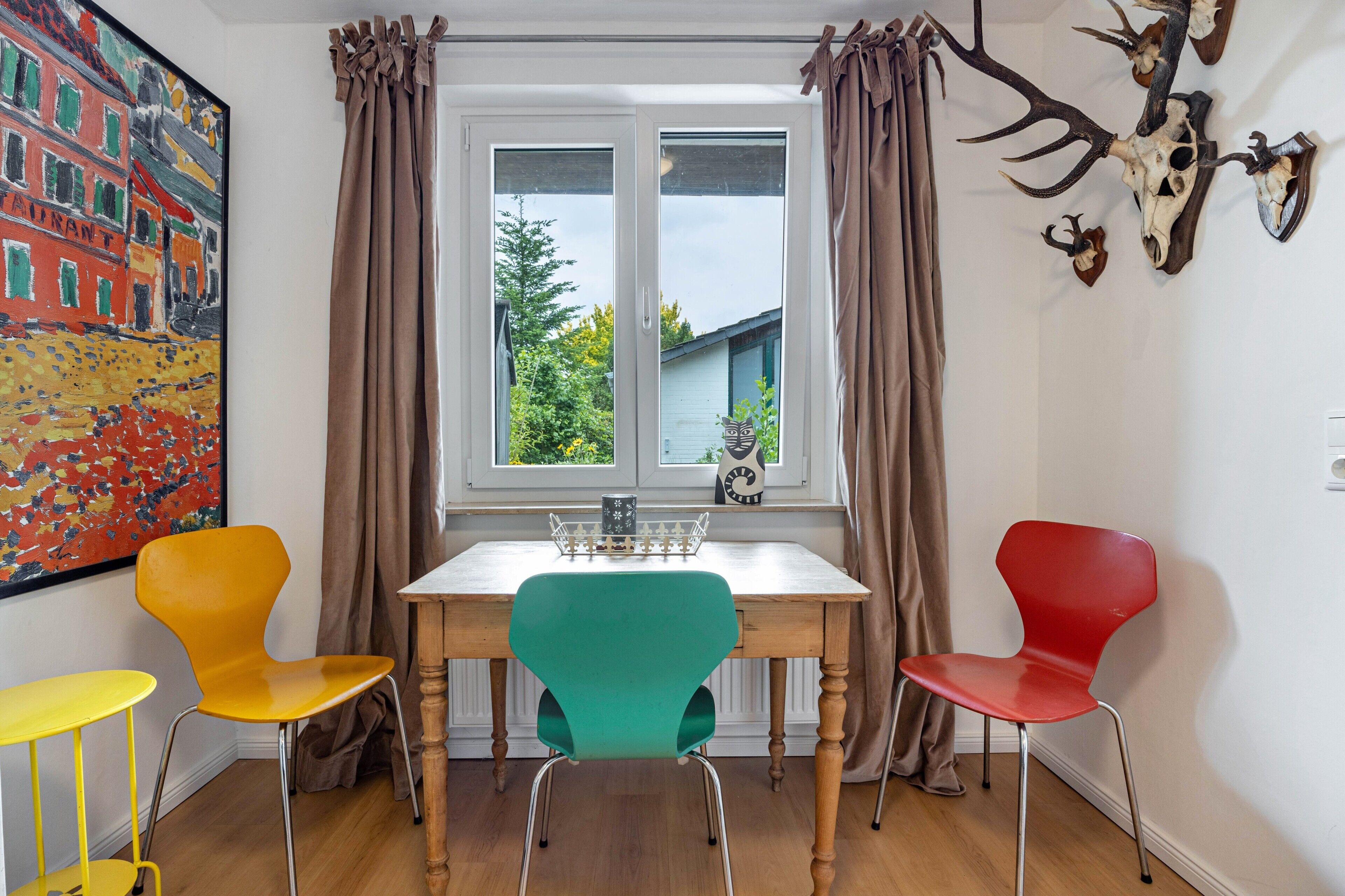 Ein Zimmer mit Esstisch, zwei Stühlen, einem Fenster mit Vorhängen, einer Wanduhr und einem Gemälde.