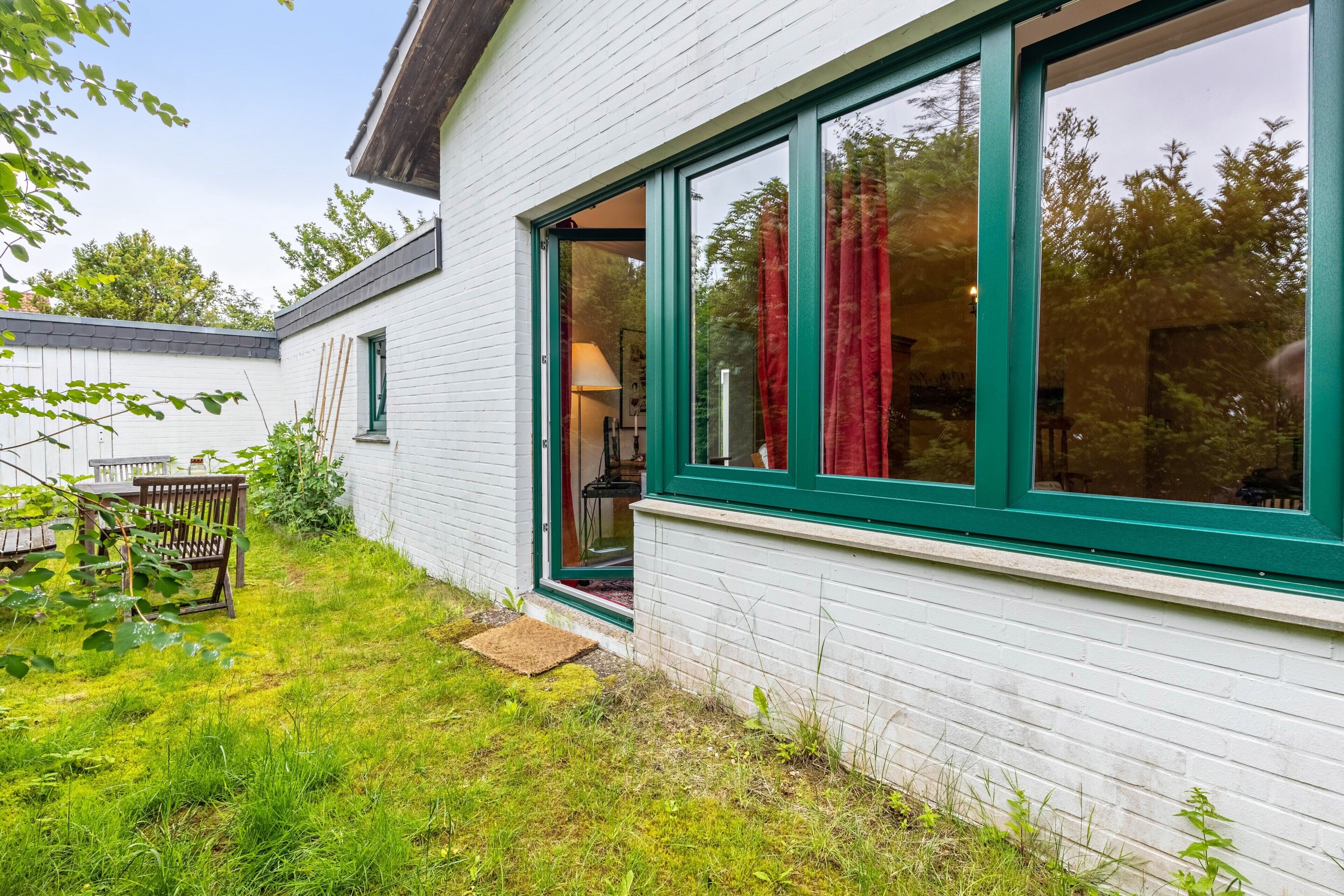 A white building with green-framed windows, a wooden bench, and a small garden area.