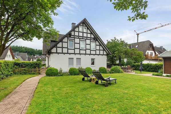 Une maison traditionnelle à colombages, avec un chemin pavé, une pelouse agrémentée de deux chaises longues et une table de pique-nique.