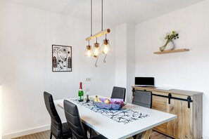 Un coin repas avec une table blanche, des chaises noires, un lustre, un panier de fruits et un meuble en bois abritant un téléviseur.