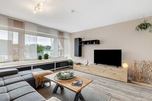 A modern living room with a sofa, a coffee table with a magazine, a TV on a wooden stand, and a window with a view of greenery. - Holiday Apartment 'Blumenau' with Private Terrace, Garden, and Wi-Fi (Schmallenberg)