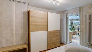 Un dormitorio con una cama, un armario de madera, un banco y vistas a una cocina y zona de comedor a través de una puerta abierta.