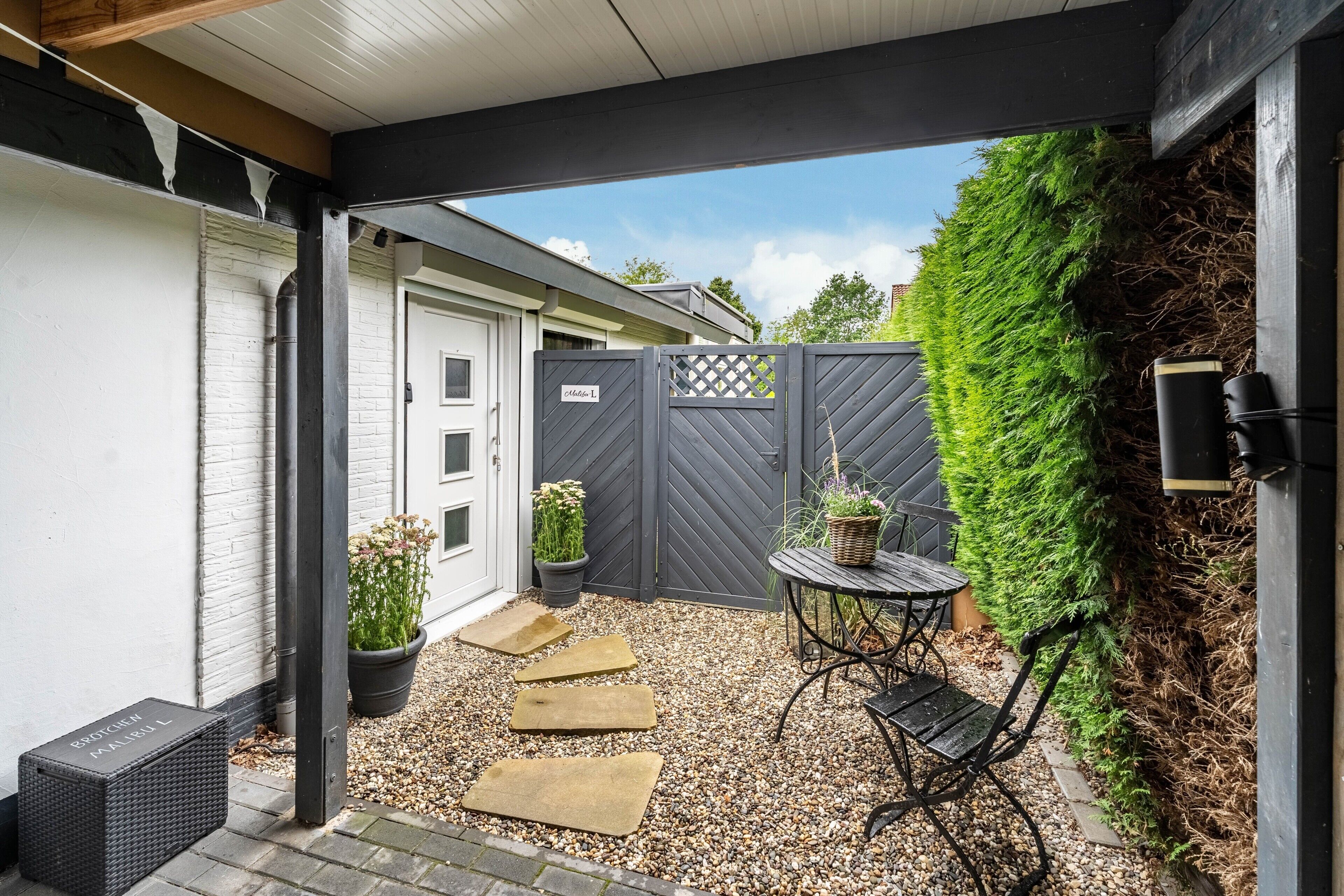 Een overdekt terras met een grindvloer, een zwarte metalen tafel en stoel, potplanten en een grijze afrastering.