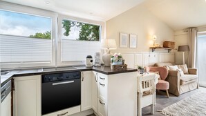 A modern kitchen with white cabinetry, a black dishwasher, and a dining area with a pink chair and a beige sofa. - Studio Malibu S by the Lake (Wettringen)