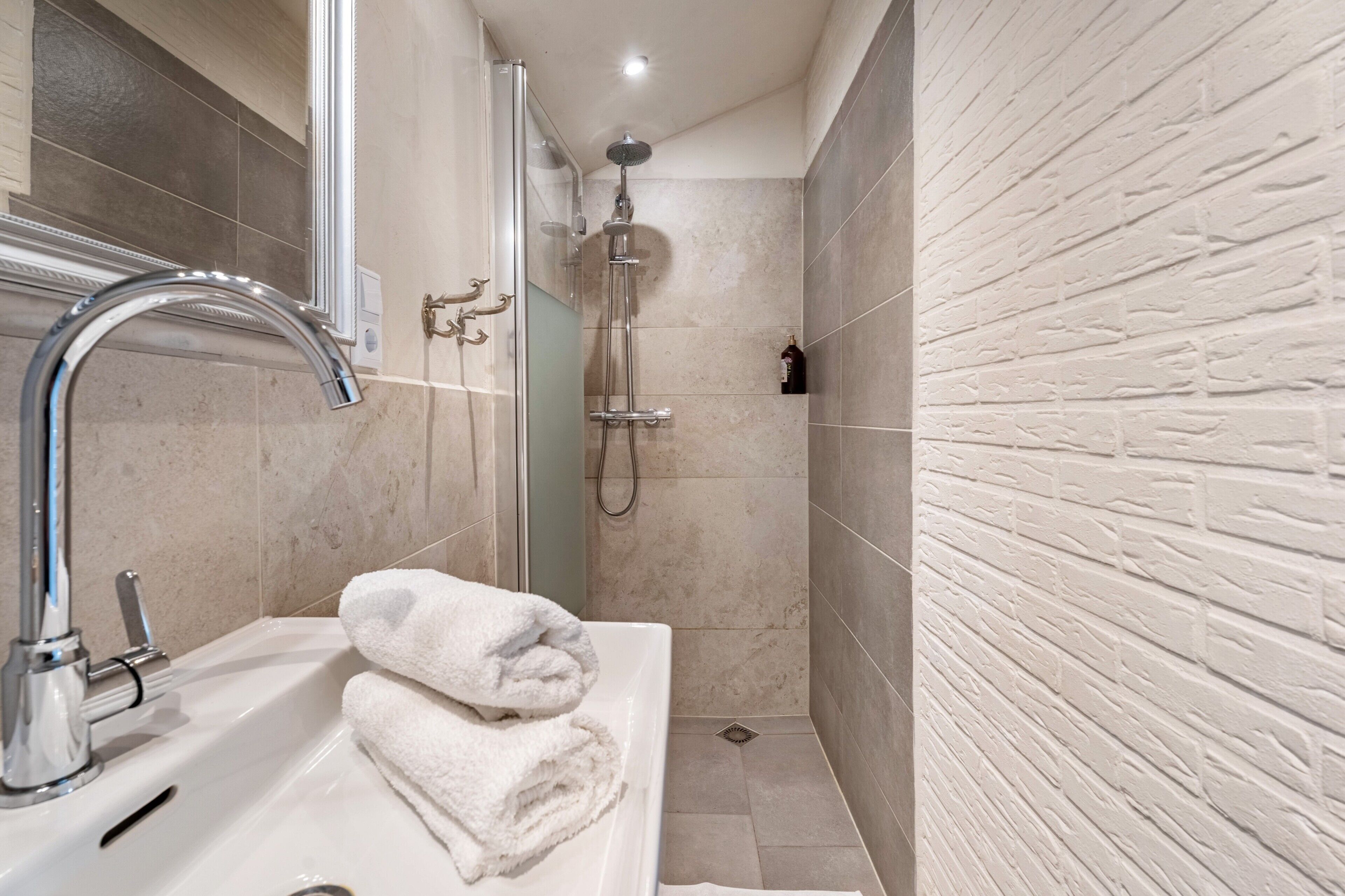 A bathroom with a sink, two towels, a shower, and a brick-patterned wall.