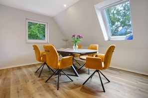 Un coin repas avec une table ronde, six chaises jaunes, un vase avec des fleurs violettes et deux fenêtres donnant sur un paysage verdoyant.