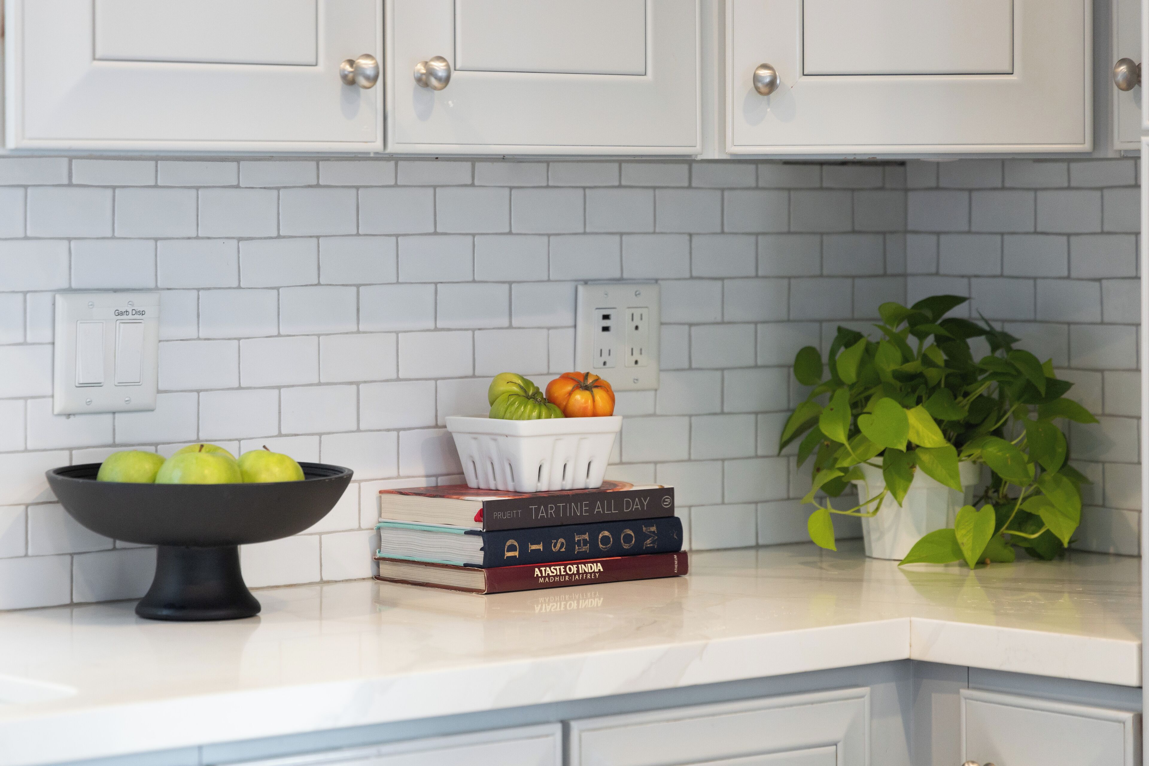 A kitchen counter with a white tiled backsplash, a bowl of green apples, a basket with a pumpkin, and a potted plant.