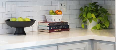 A kitchen counter with a white tiled backsplash, a bowl of green apples, a basket with a pumpkin, and a potted plant.