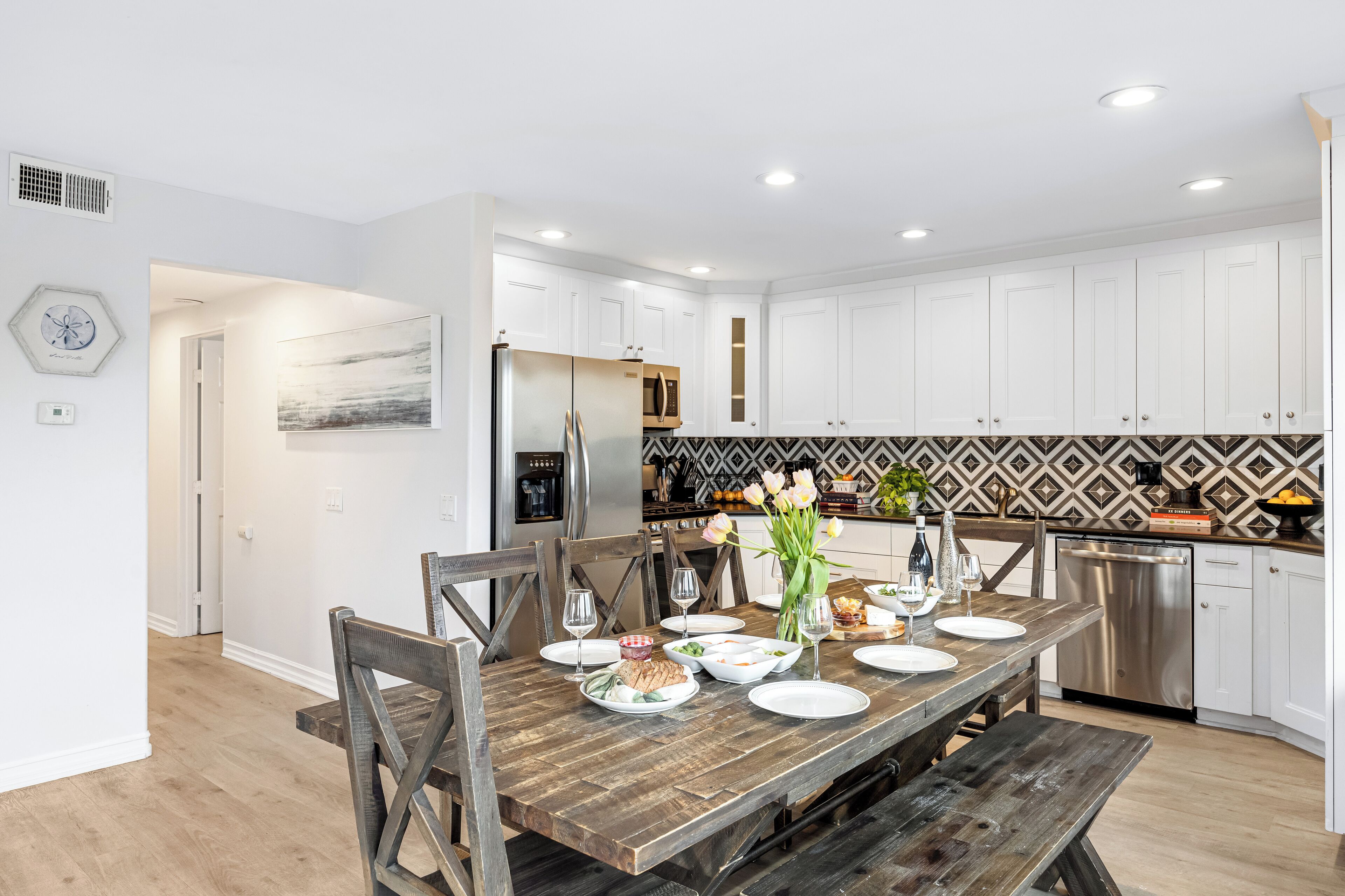 Une cuisine moderne avec des meubles blancs, une table à manger en bois pour six personnes et un revêtement mural décoratif à motifs géométriques.