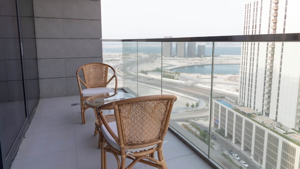 Un balcón con sillas de mimbre y una mesa de vidrio, con vistas a un paisaje urbano que incluye rascacielos y un puerto deportivo.