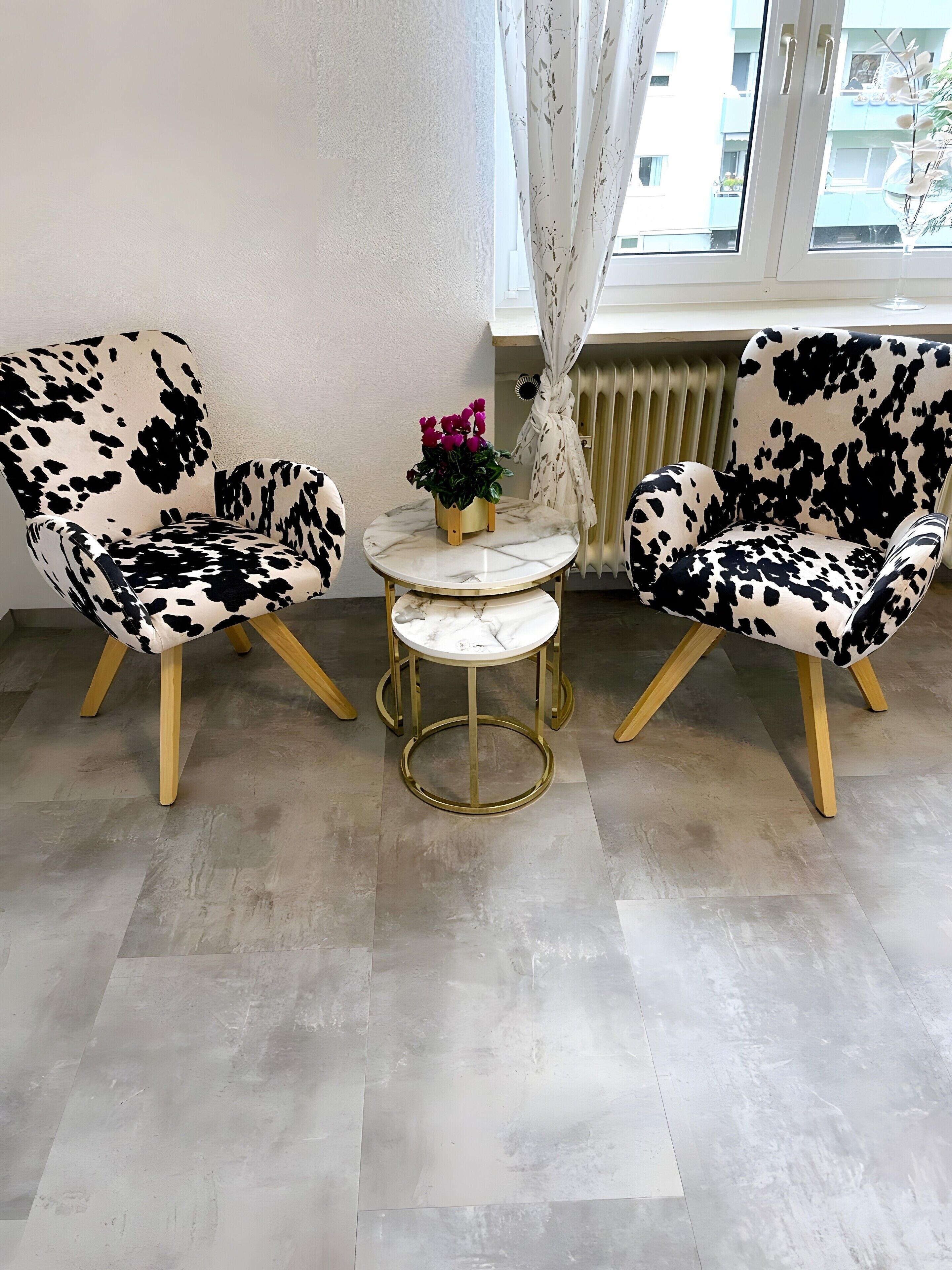 A room with two black and white patterned chairs, a small round table with a gold base, and a potted plant with pink flowers.