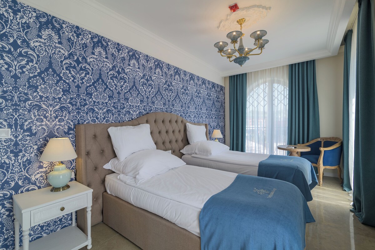 A hotel room with a tufted headboard bed, a bedside table with a lamp, a chair, and a view of the outdoors through a window with curtains.