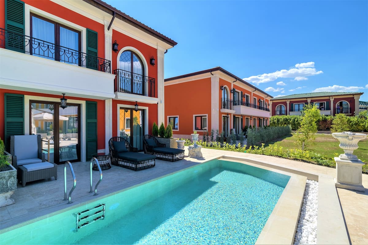 A pool area with lounge chairs and a clear blue pool, surrounded by a well-maintained garden and a red building with balconies.