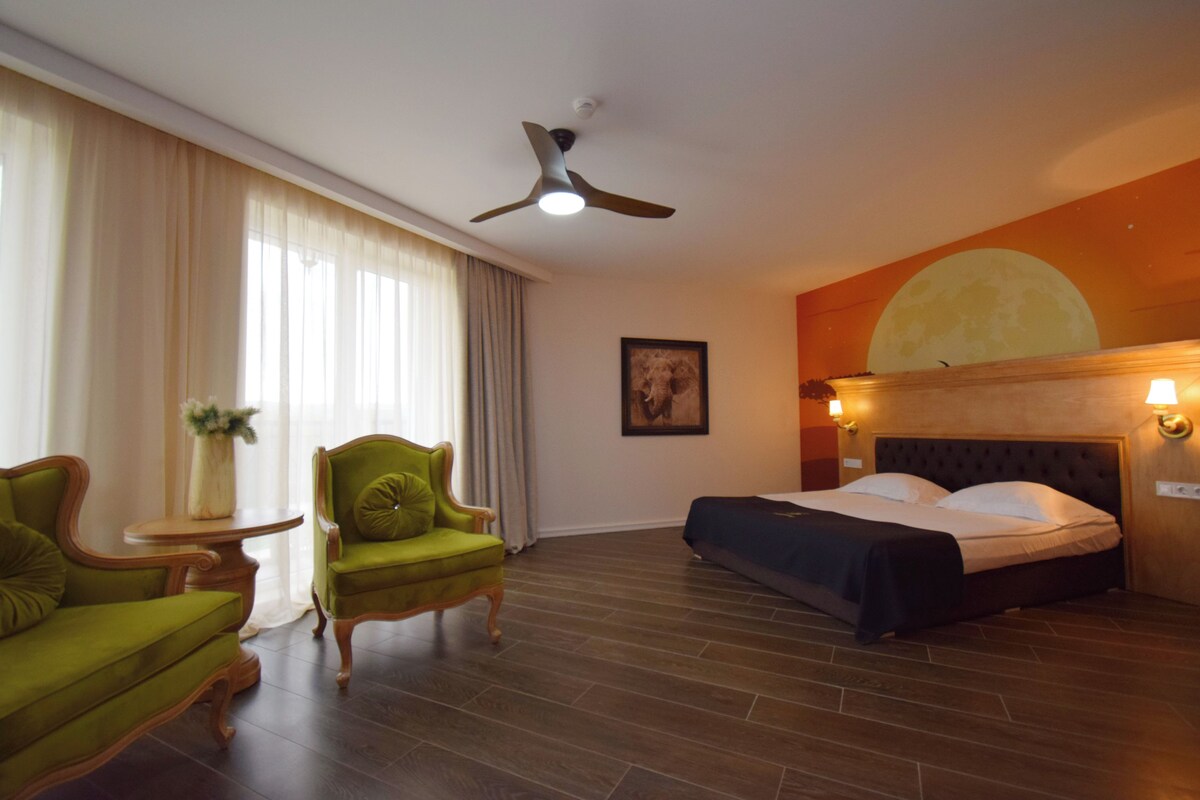 A hotel room with a bed, two green armchairs, a small table with a vase, a ceiling fan, and a large wall mural.