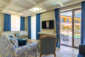 Ein Wohnzimmer mit Sofa, Sessel und Fernseher. Ein Balkon mit Gartenmöbeln ist sichtbar.