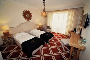 A hotel room with two beds, a desk, a chair, a TV, and a large window with curtains. - Eco Therma Village Spa & Sport (Balchik)