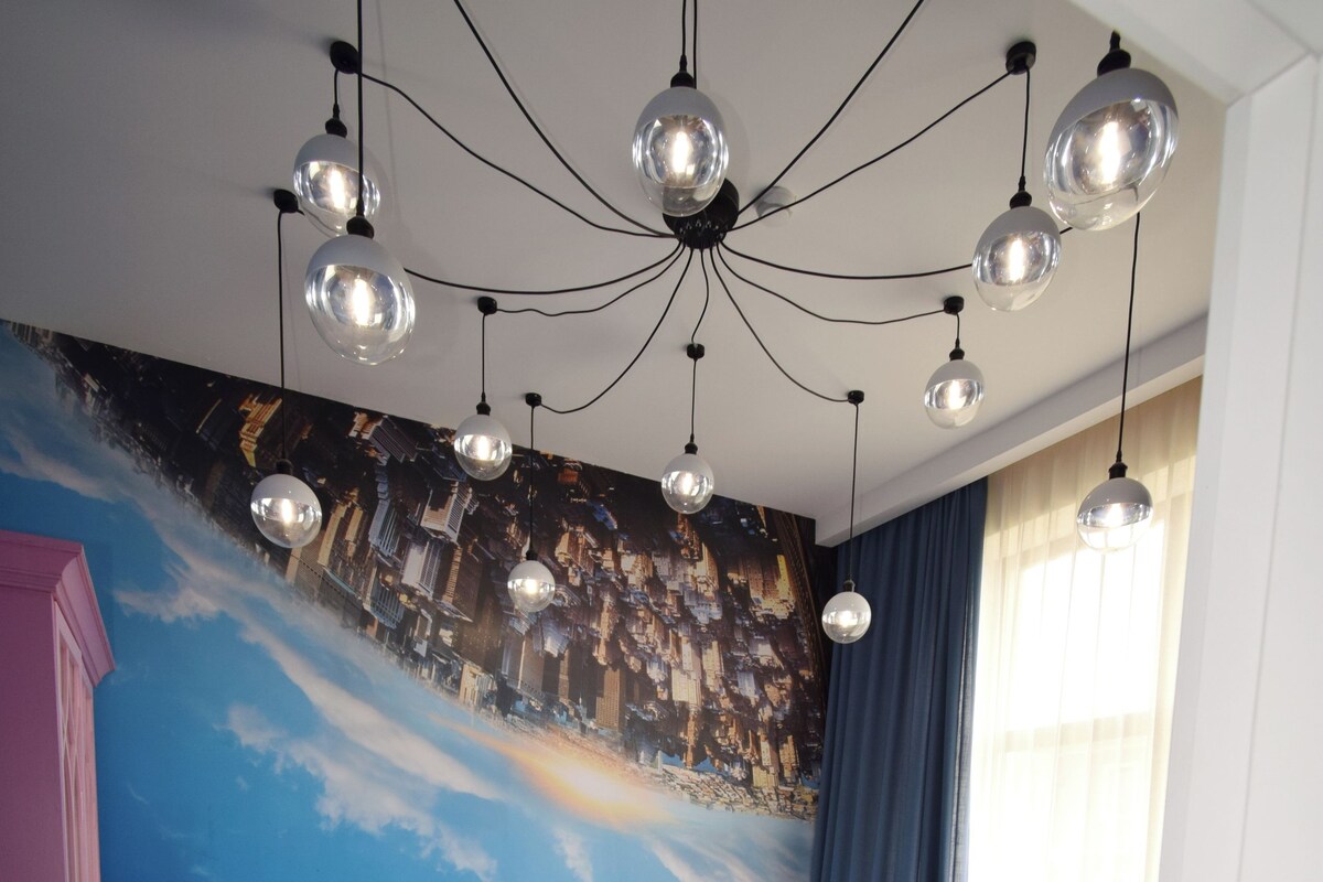 A bedroom with a bed, a dresser, a wall mural, and a ceiling light fixture.