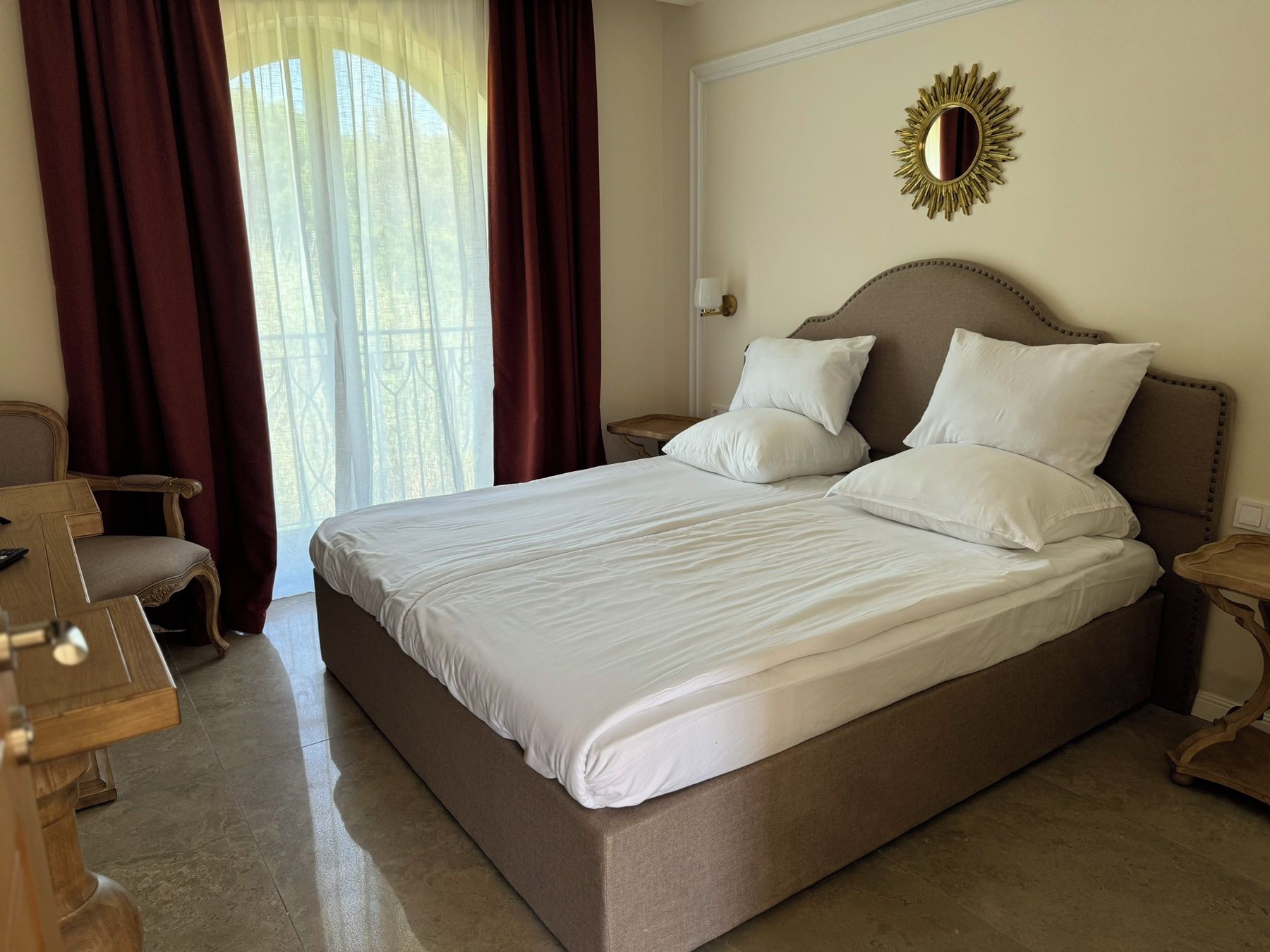 Una cama grande con sábanas blancas, una mesita de noche de madera, una silla y una ventana con cortinas.
