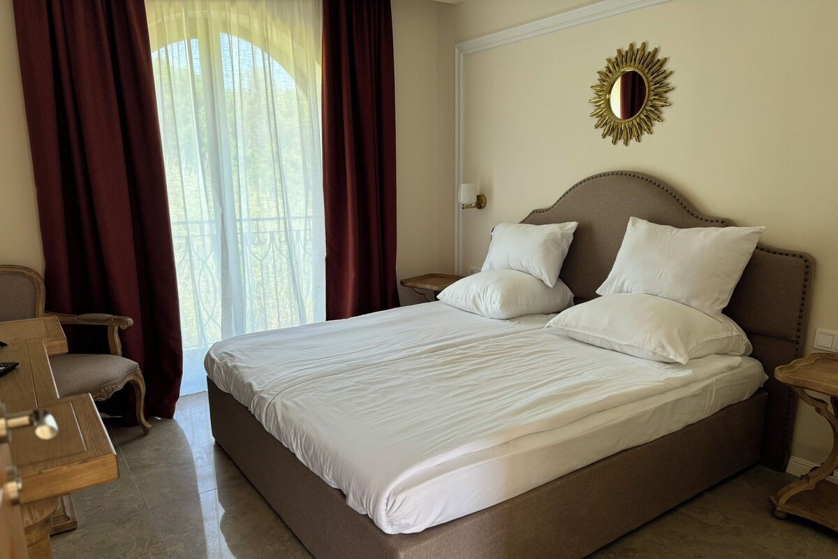 A large bed with white linens, a wooden nightstand, a chair, and a window with curtains.