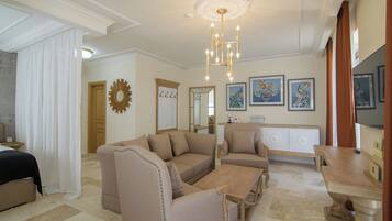 A spacious living room with a large beige sofa, a smaller armchair, a wooden coffee table, and a dining table with chairs. There is a chandelier hanging from the ceiling, a large mirror on the wall, and framed artwork above the fireplace.