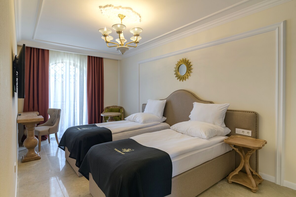 A hotel room with two beds, a desk, a chair, a TV, and a chandelier.