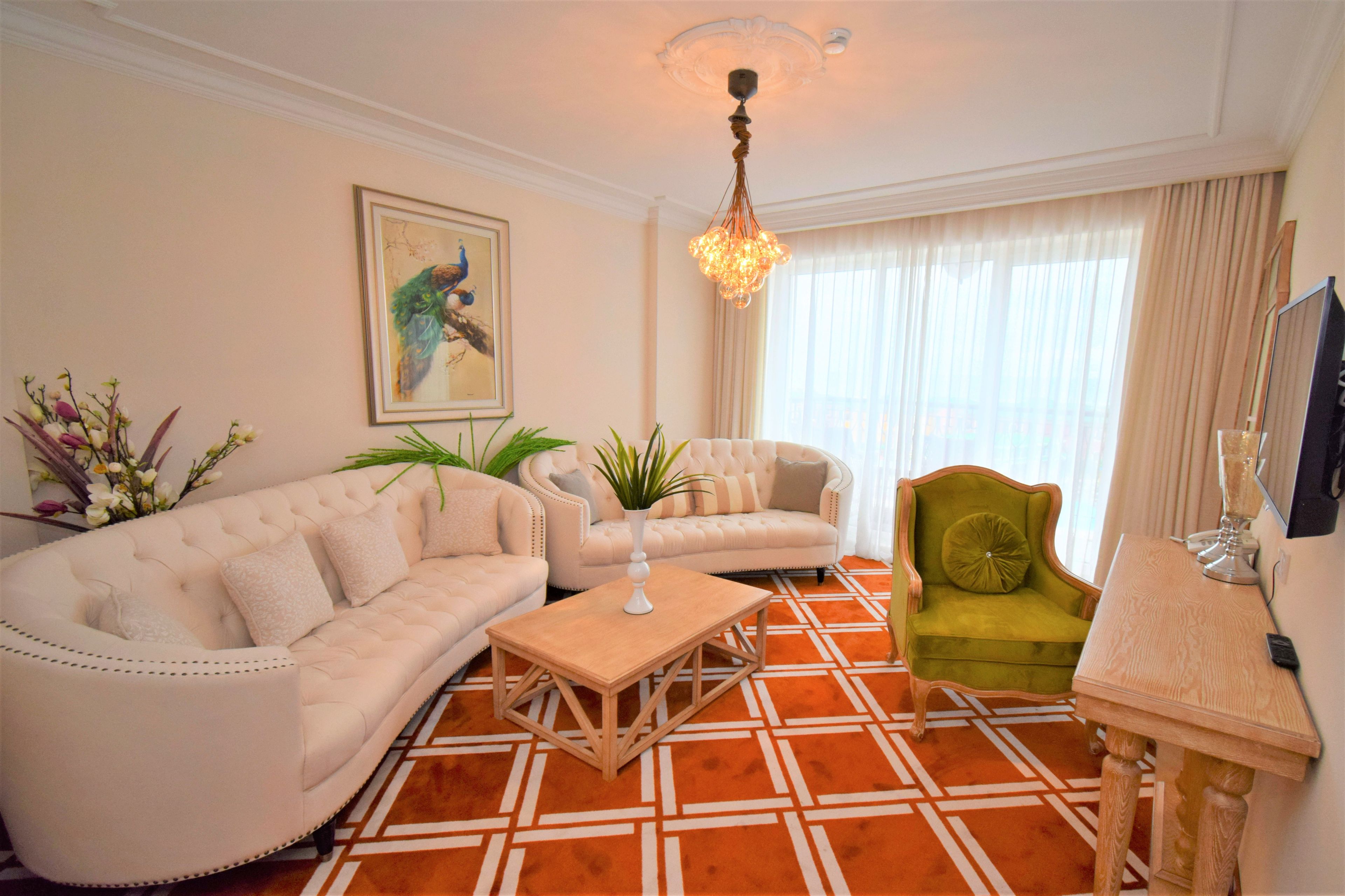 A living room with a beige sofa, a green armchair, and a wooden coffee table. There is a painting on the wall and a chandelier hanging from the ceiling.