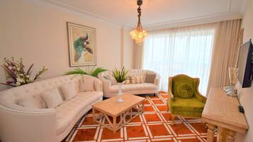 A living room with a beige sofa, a green armchair, and a wooden coffee table. There is a painting on the wall and a chandelier hanging from the ceiling.