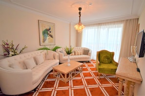 Una sala de estar con un sofá color beige, un sillón verde y una mesa de centro de madera. Hay un cuadro en la pared y una araña colgando del techo.
