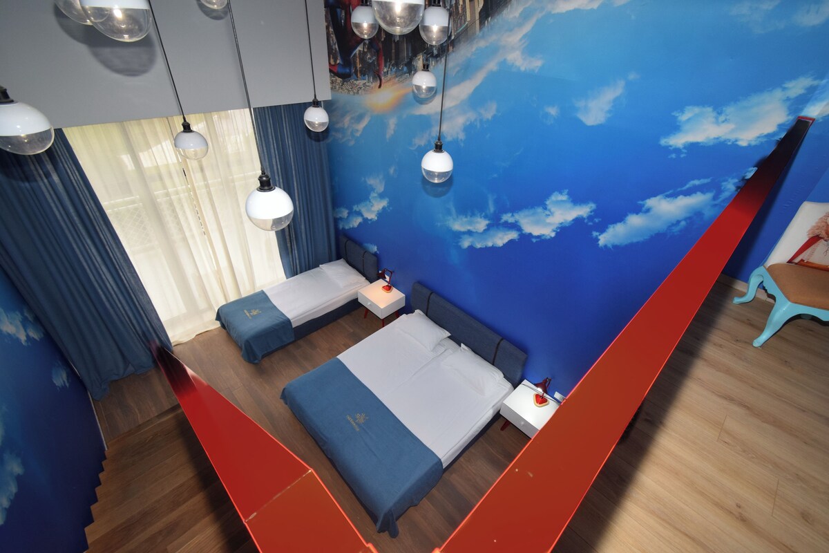 A modern hotel room with a large bed, a desk, and a seating area. There are multiple light fixtures and a mural on the wall.