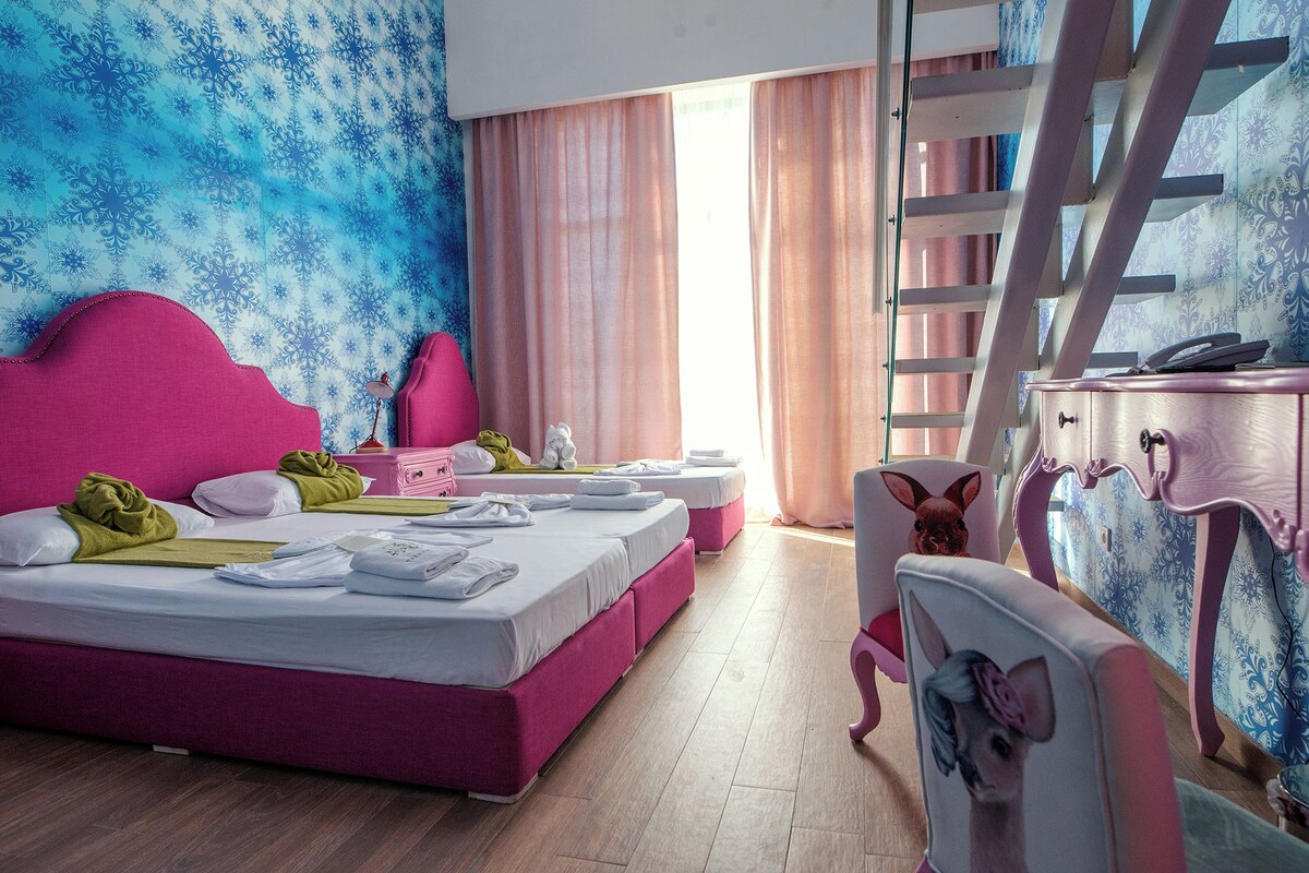 A hotel room with a pink bed, a desk, and a chair. There is a window with curtains and a ladder.