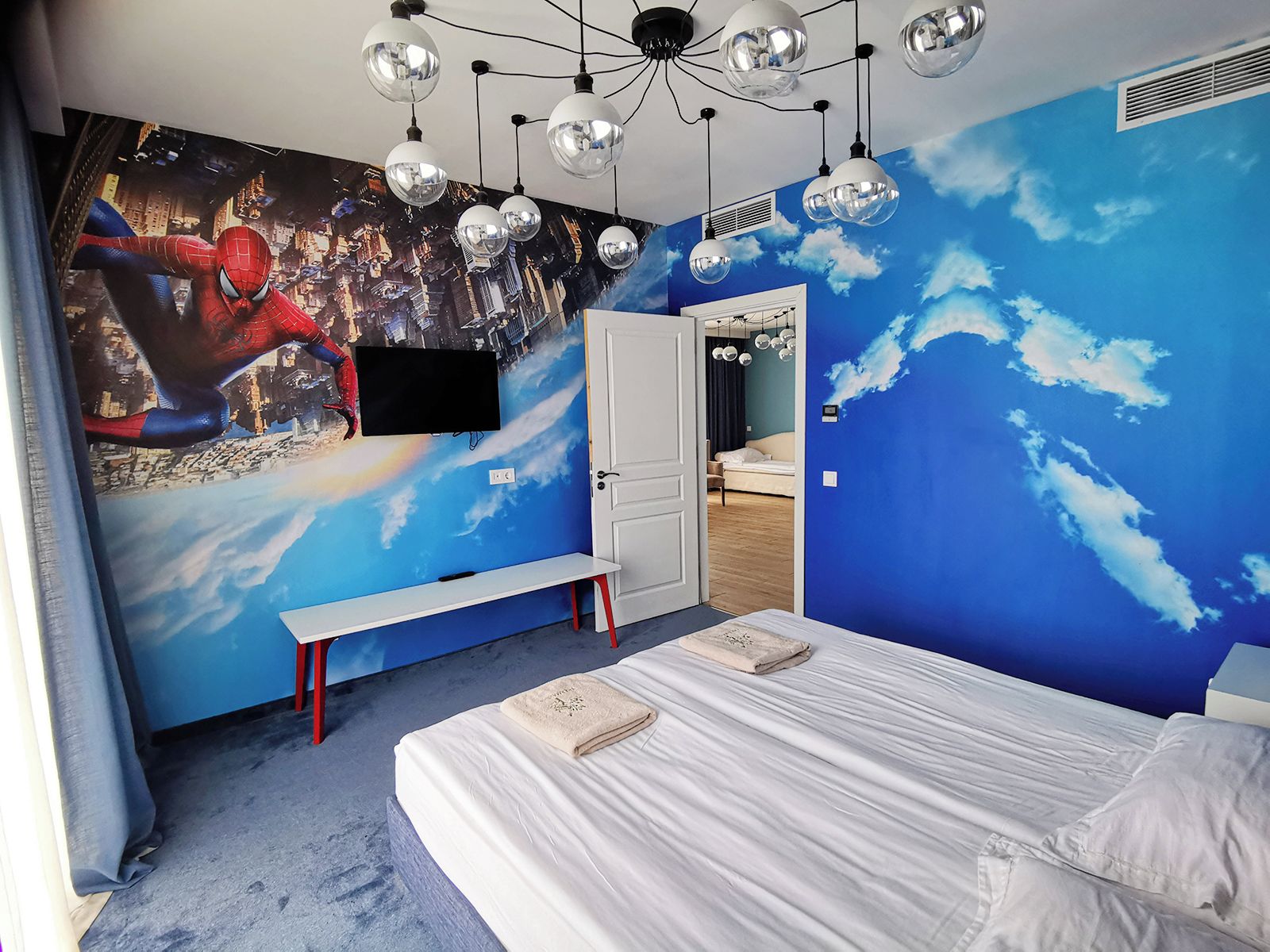 Un dormitorio con un mural de un paisaje urbano y nubes, una cama, un banco y un armario.