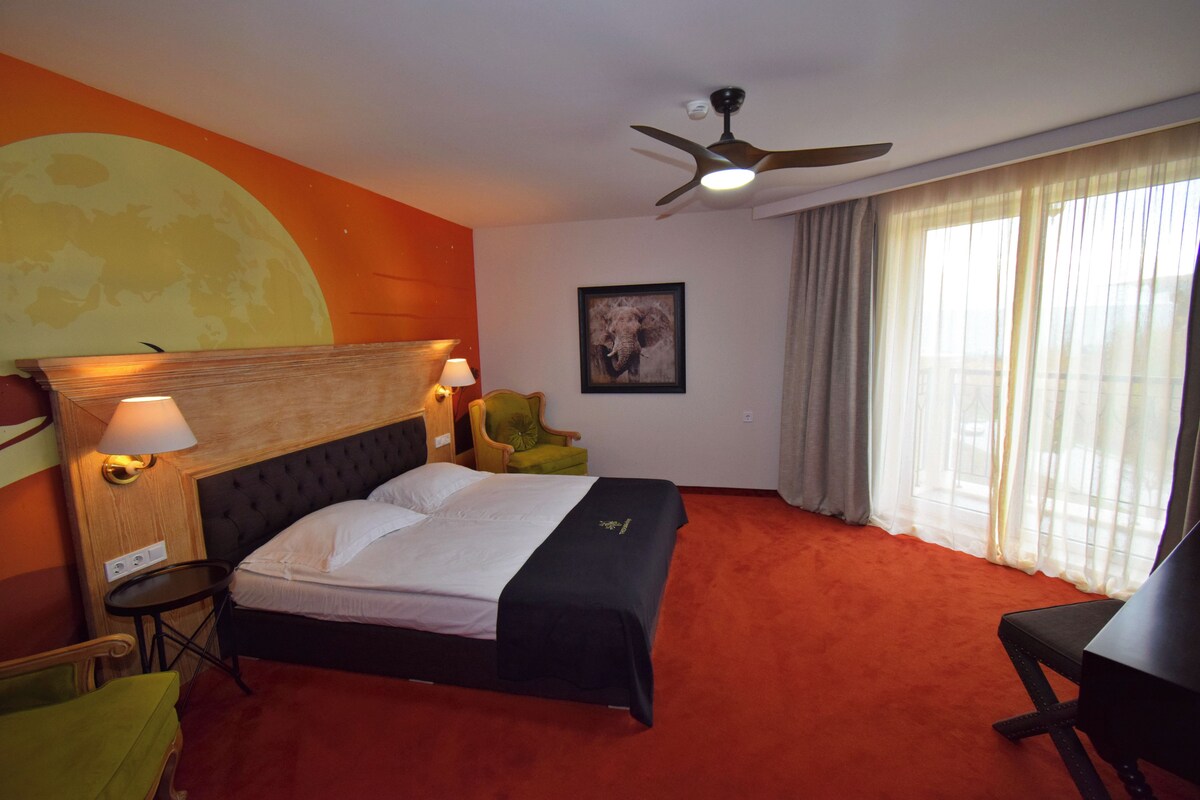 A hotel room with a bed, a desk, a chair, a lamp, a ceiling fan, and a large window with curtains.