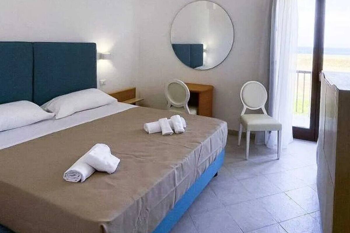 A hotel room with a bed, a round mirror, a chair, and a view of the outdoors.