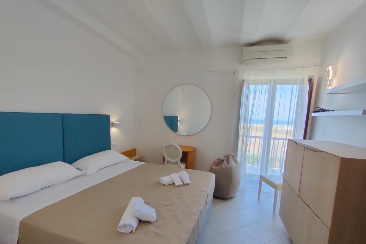 Ein Hotelzimmer mit einem Bett, einem runden Spiegel, einem Sessel, einem Schreibtisch und Blick ins Freie durch ein Fenster.