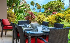 An outdoor dining area with wicker chairs and red cushions, surrounded by tropical plants and palm trees.