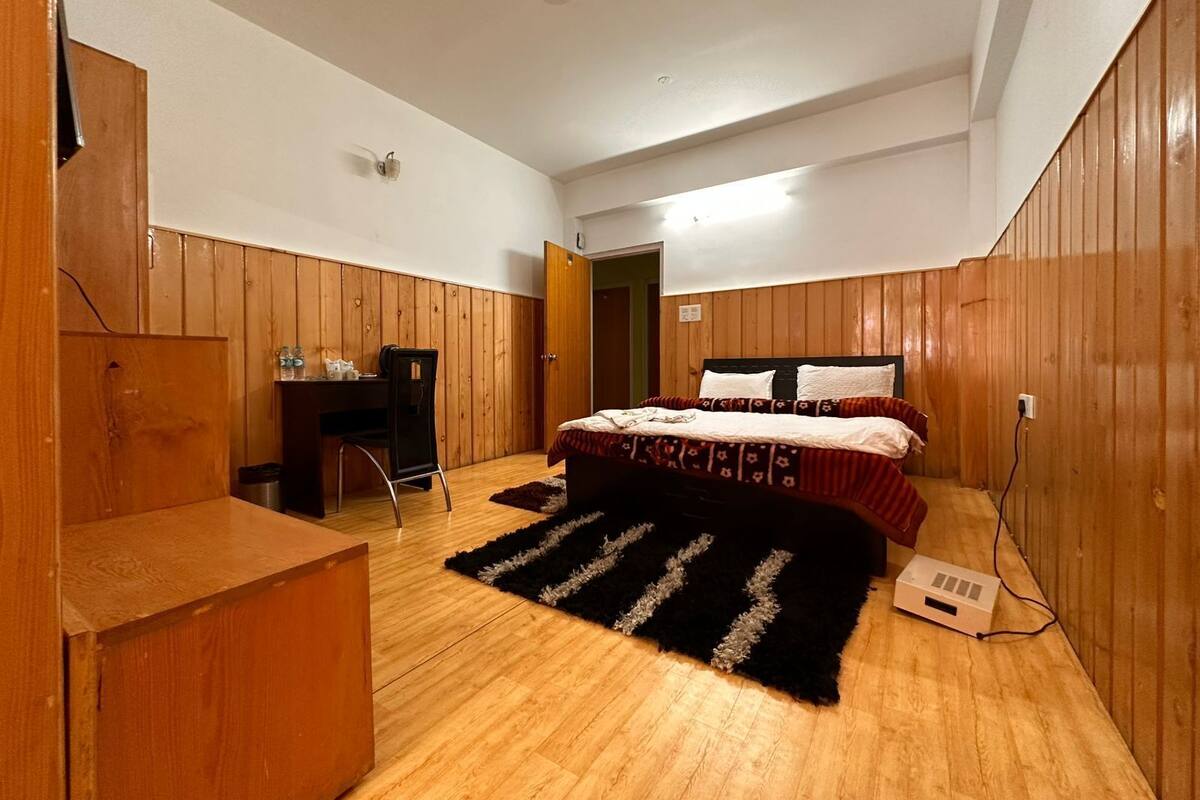 A bedroom with a wooden floor, a bed with patterned bedding, a desk with a chair, and a black rug.