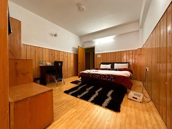 A bedroom with a wooden floor, a bed with patterned bedding, a desk with a chair, and a black rug. - Hotel Tashi Ga-Tsel (Tawang)