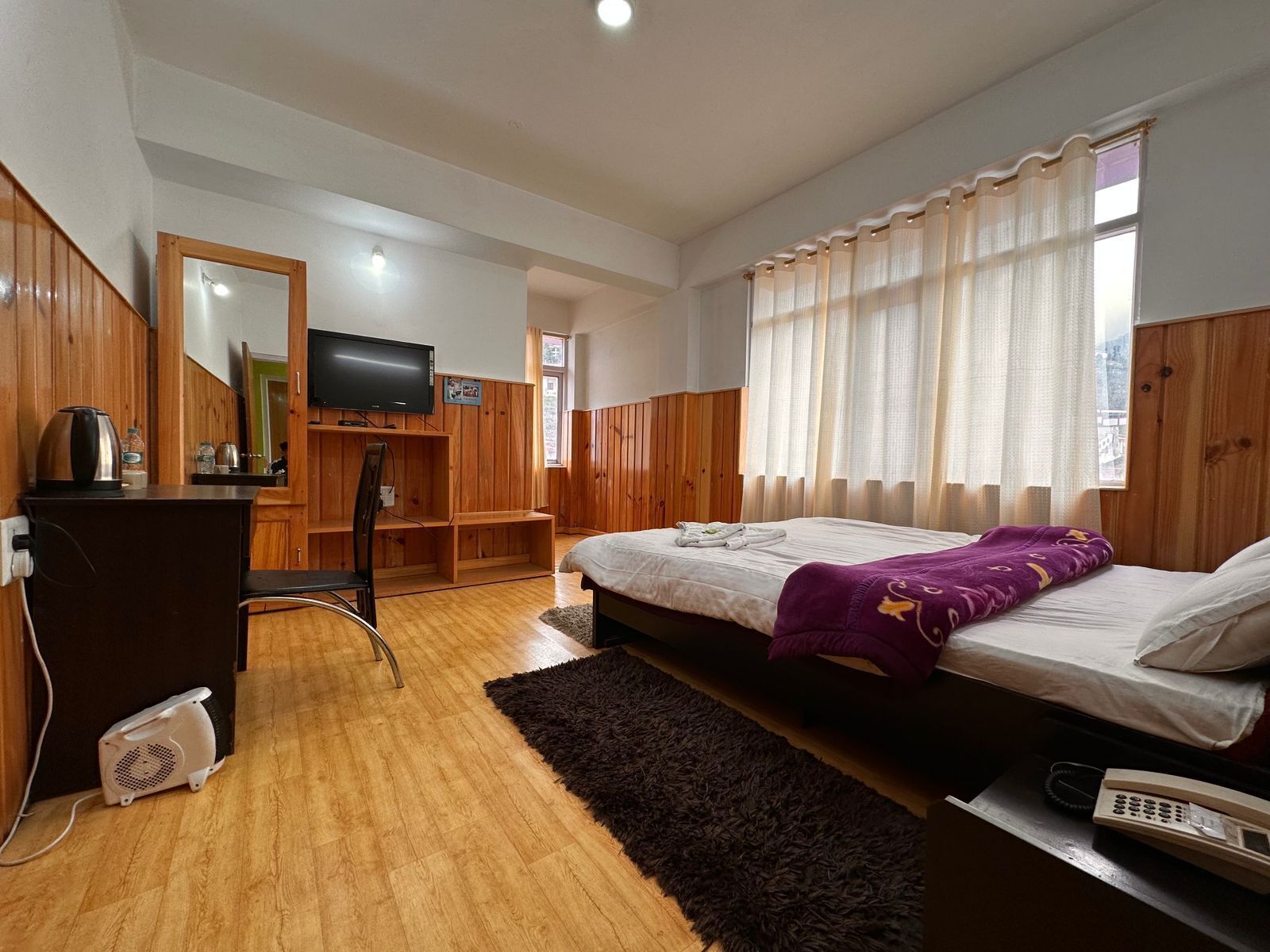 Ein Schlafzimmer mit Bett, Schreibtisch, Stuhl, Fernseher und einem Fenster mit Vorhängen.