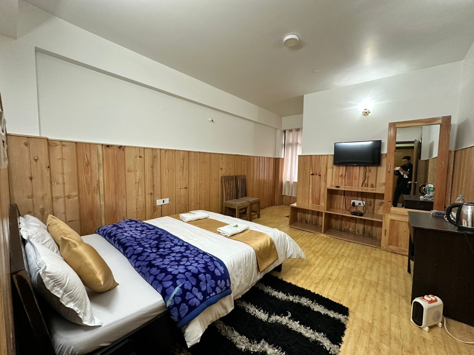 Een hotelkamer met een wand van houten panelen, een bed met wit en gouden beddengoed, een aan de muur bevestigde televisie, een bureau met stoel, en een kleine verwarming op de vloer.