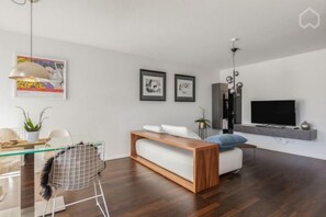 Een moderne woonkamer met een glazen eettafel, een bank, een televisie en kunstwerken aan de wand.