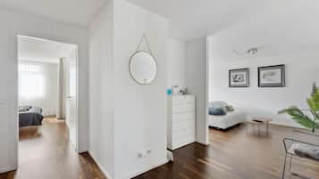 Ein modernes, helles Zimmer mit Holzboden, einem weißen Kleiderschrank, einem runden Spiegel, einem kleinen Tisch und einer Couch. An den Wänden befinden sich gerahmte Bilder und in der Ecke steht eine Pflanze.