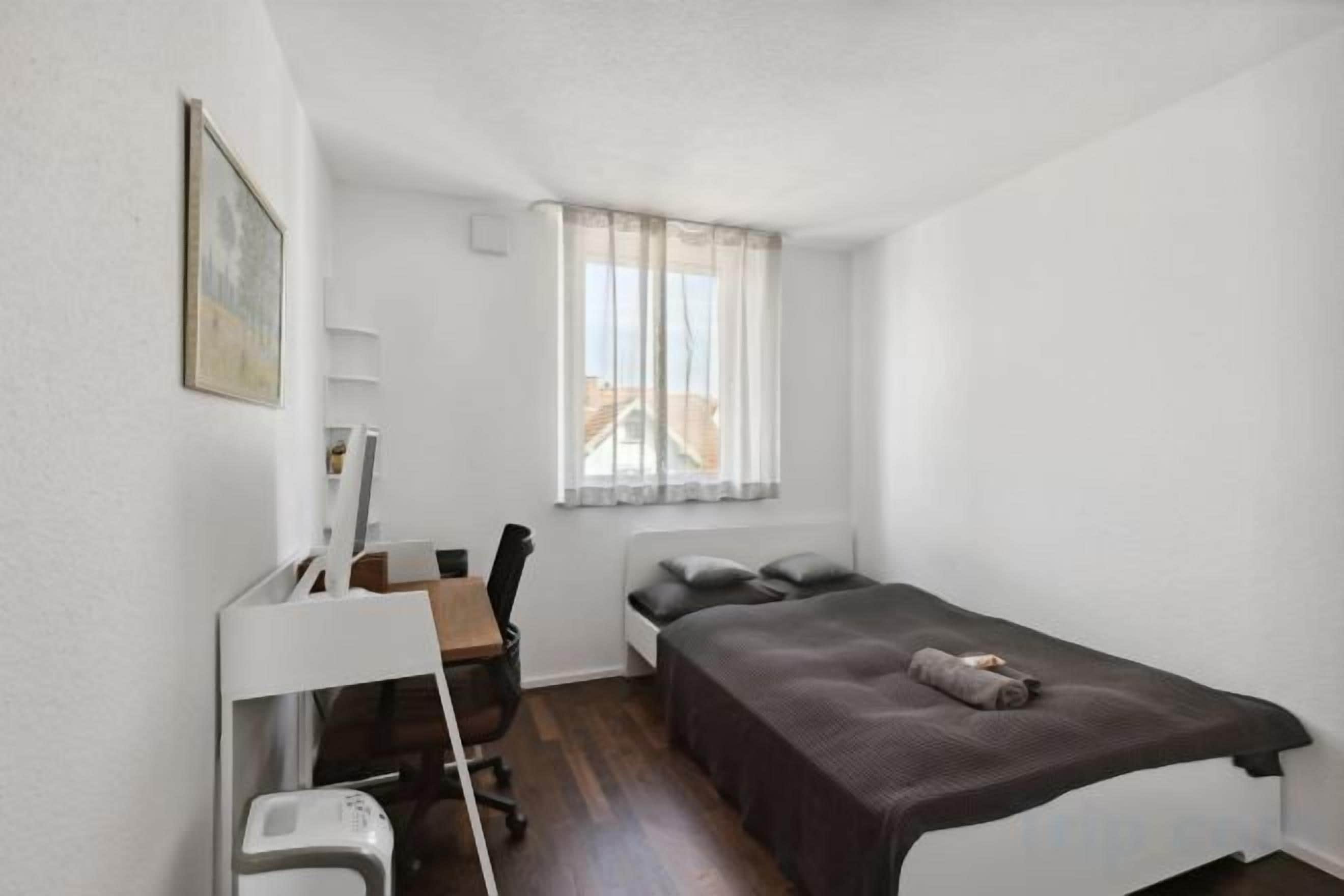 Eine kleine, einfache Unterkunft mit Bett, Schreibtisch, Stuhl und einem blickdichten Fenster.